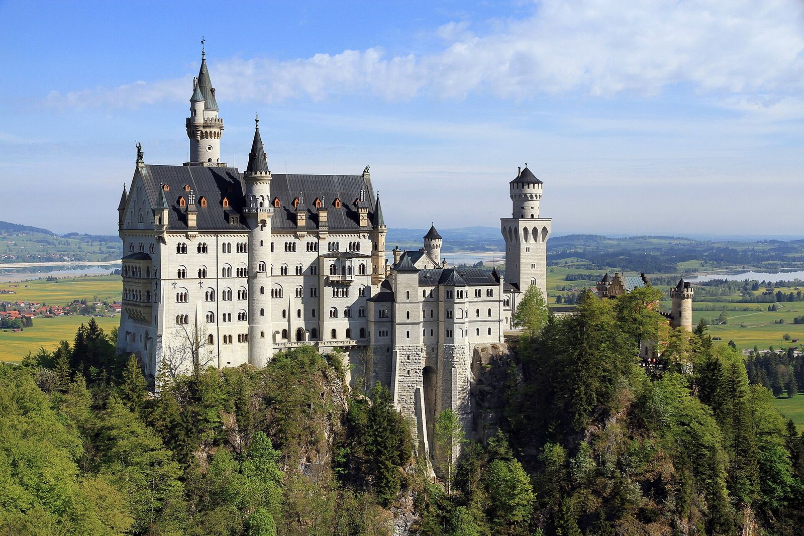 Schloss Neuschwanstein auf seinem bewaldeten Felsen, im Hintergrund das Alpenvorland mit Seen, gesehen von der Marienbrücke
