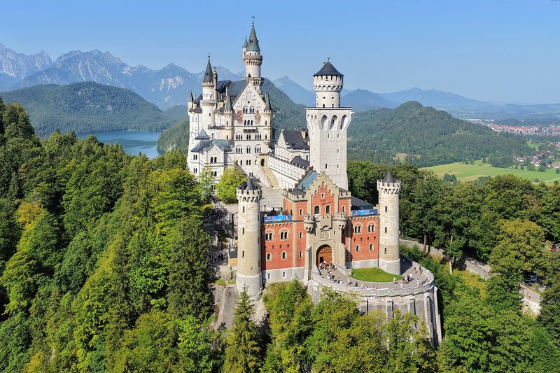 Schloss Neuschwanstein von Osten mit dem rötlichen Torbau im Vordergrund, dahinter Alpsee und Berggipfel